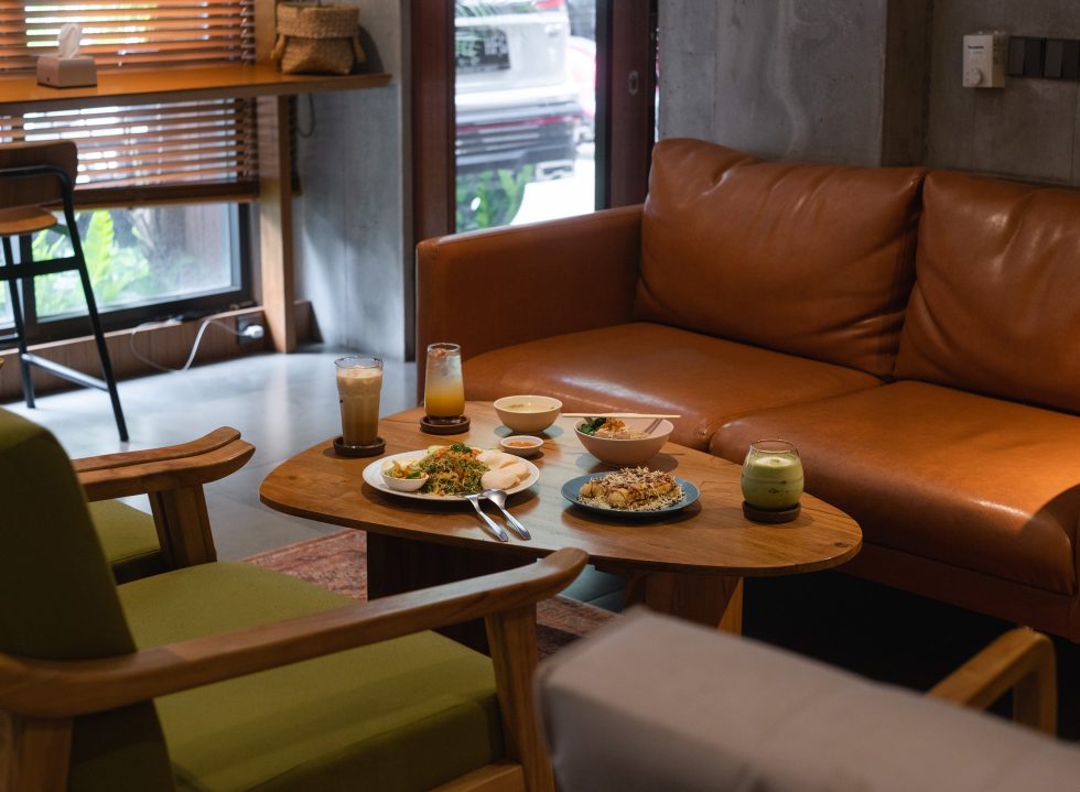 Meja kayu berisi nasi goreng, lauk pendamping, es kopi susu, dan matcha di depan sofa kulit cognac